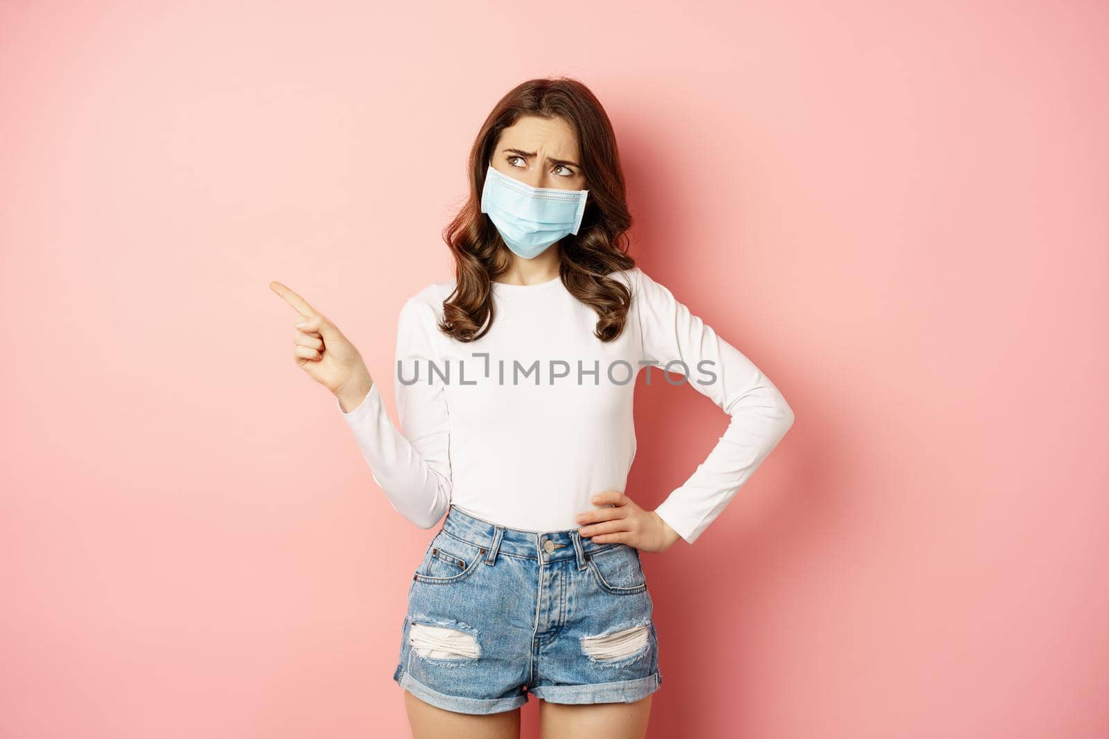 Confused, indecisive young woman in medical face mask, pointing finger left and looking complicated, making decision, standing over pink background by Benzoix