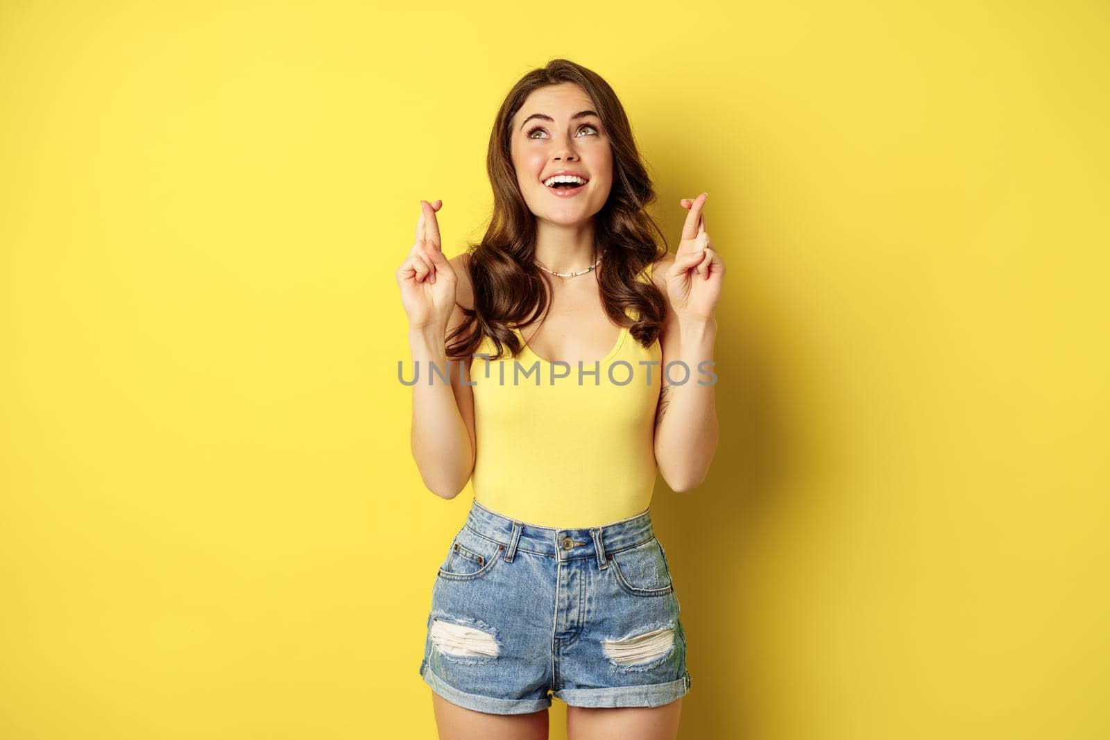 Hopeful young beautiful woman praying, believe, hoping to receive smth, cross fingers for good luck, anticipating, standing over yellow background by Benzoix