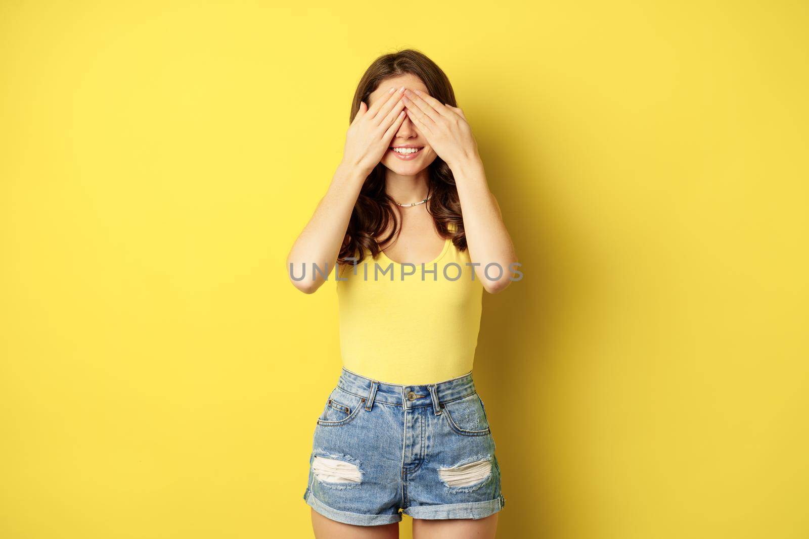 Portrait of brunette woman cover eyes with hands, blindfolded, smiling with anticipation, expecting smth, waiting for surprise, standing over yellow background by Benzoix