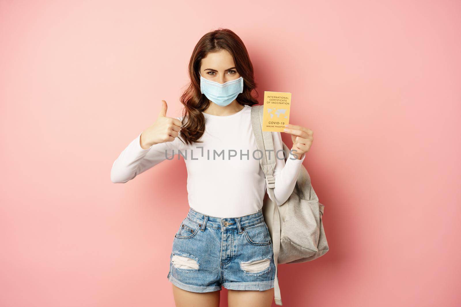 Young woman in medical mask, showing thumbs up and covid vaccination certificate, travel during pandemic, going on holiday, standing over pink background.