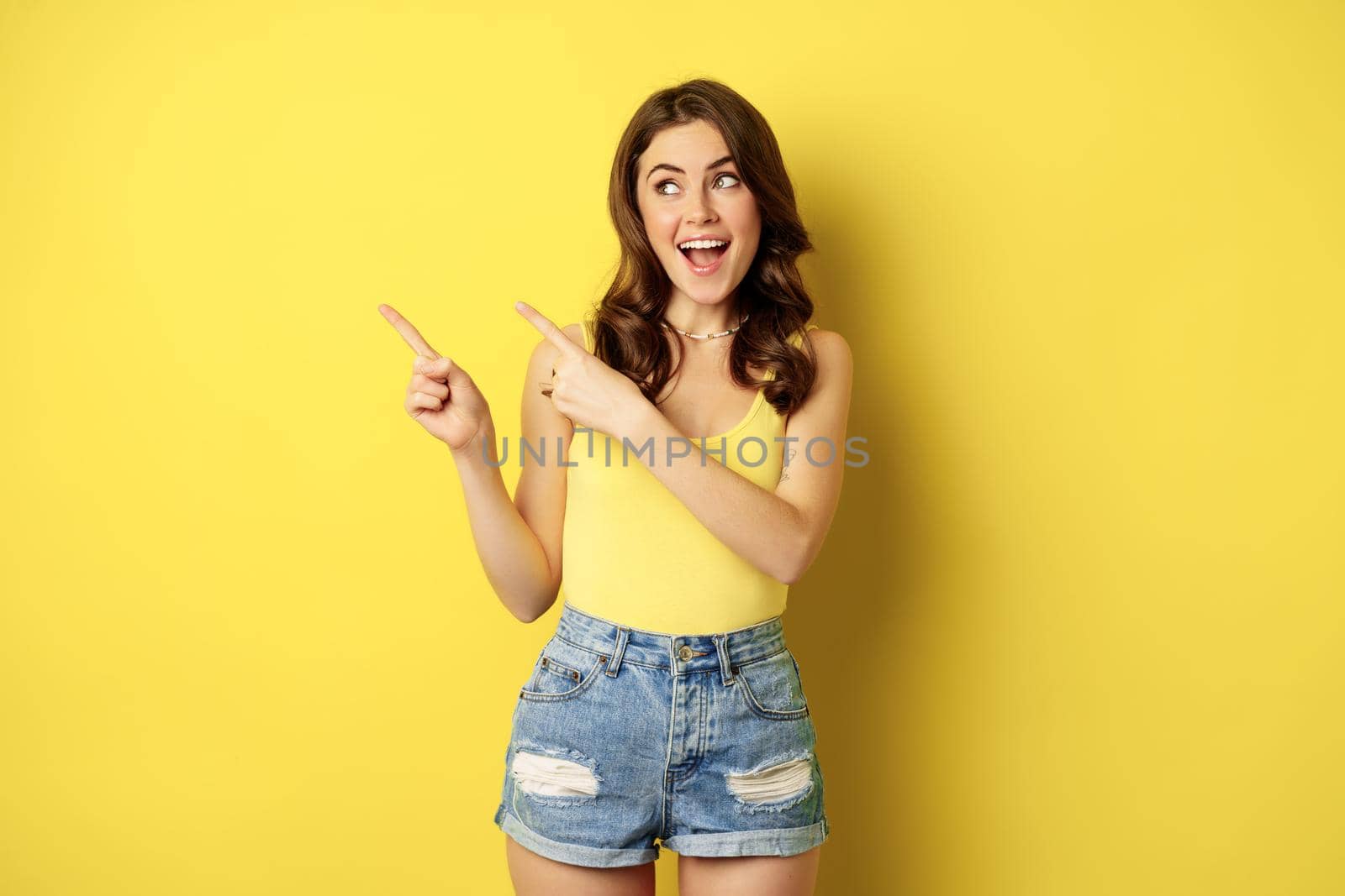 Portrait of stylish summer girl, brunette woman showing way, pointing fingers left, advertising, standing over yellow background. Copy space