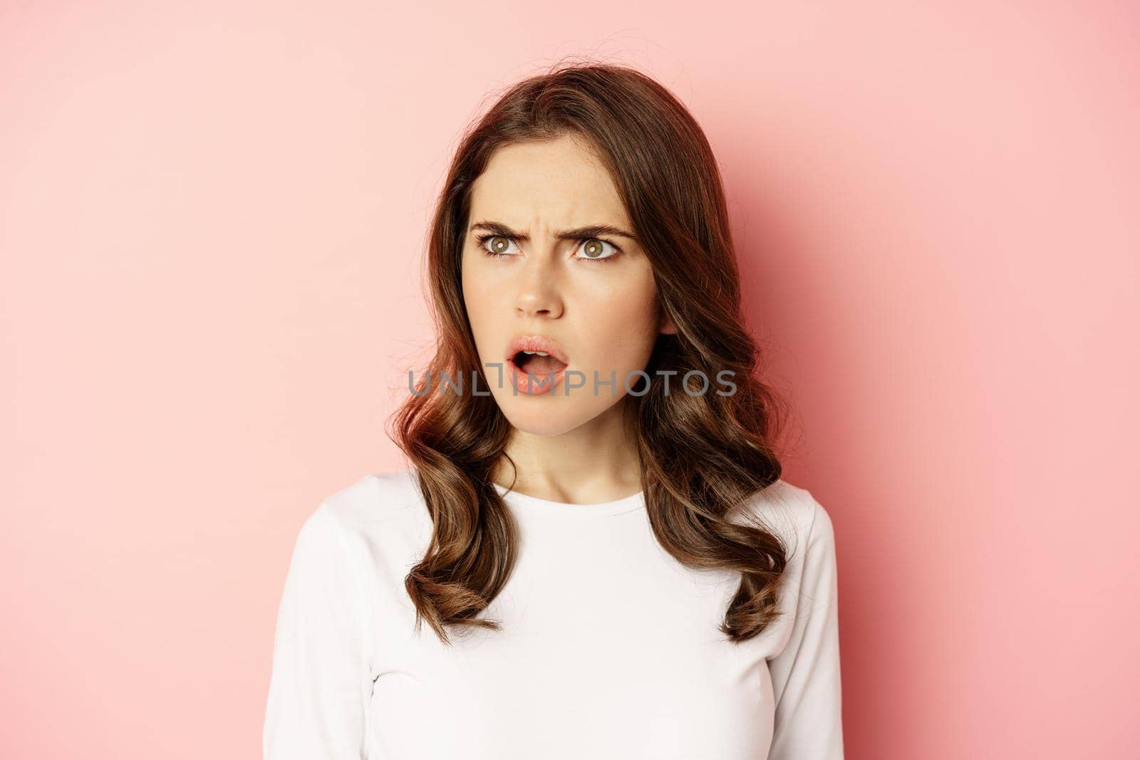 Close up portrait of confused, frustrated young woman, looking left with disappointment, puzzled by something, standing over pink background.