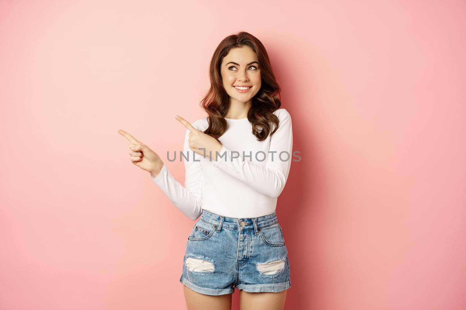 Cute smiling brunette girl, showing advertisement, pointing fingers left at banner, logo on empty pink space, studio background.