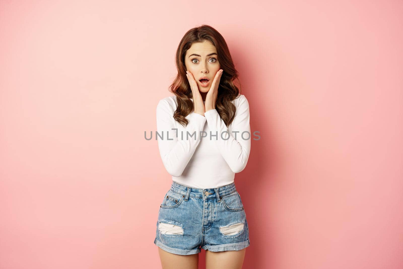 Shocked glamour girl looking at camera, gasping amazed. Woman staring surprised at advertisement, store discounts, standing over pink background.