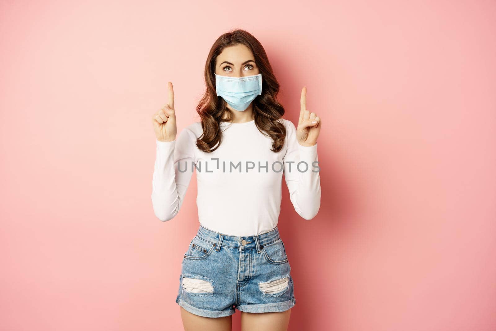 Portrait of young glamour girl in medical face mask, reading promo text, pointing and looking up, standing over pink background. Covid and pandemic concept