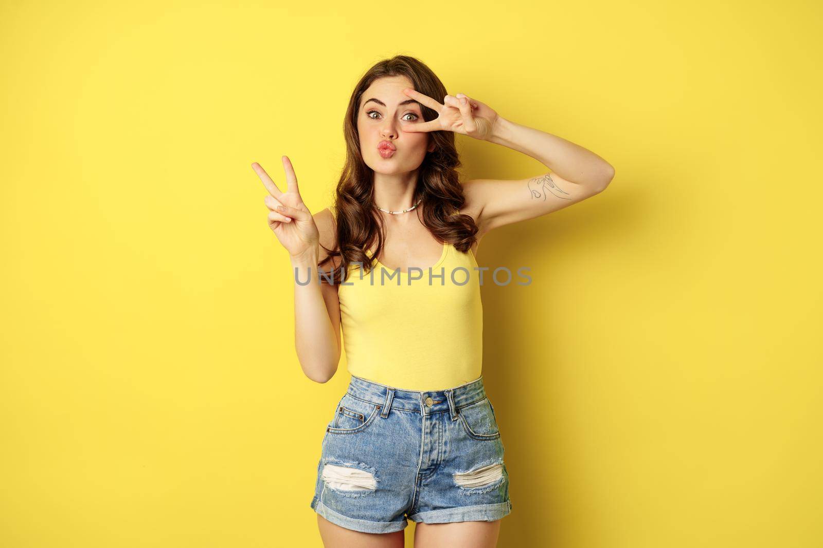 Positive girl, female model showing peace, v-sign gesture and smiling, standing in tank top and denim shorts, yellow background.