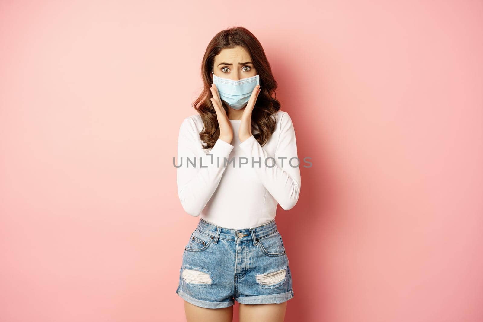 Covid-19 and quarantine concept. Stylish girl in medical face mask, looking worried and shocked at camera, standing against pink background.