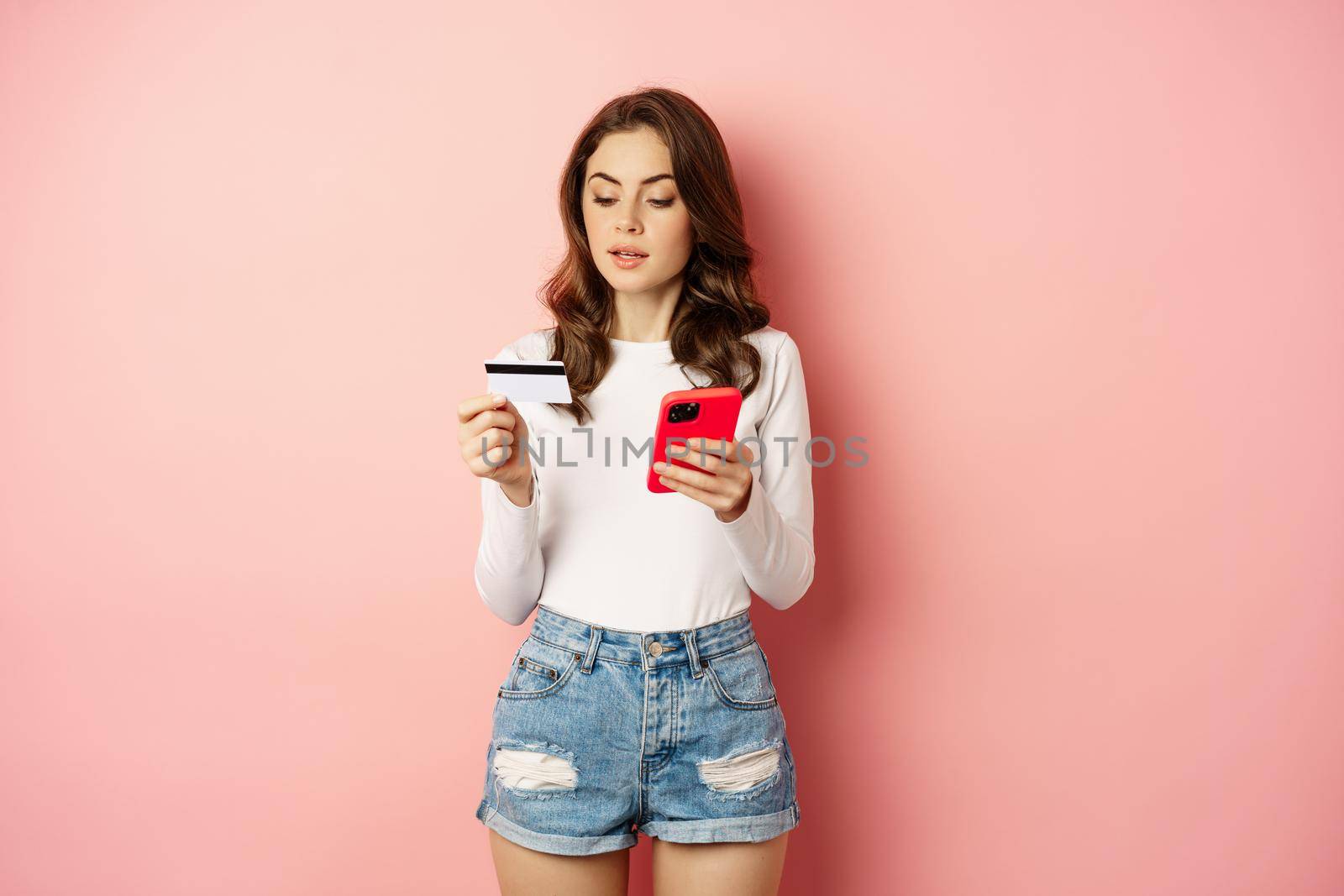 Online shopping. Young woman order online, making purchase in app, holding smartphone and credit card, standing against pink background.
