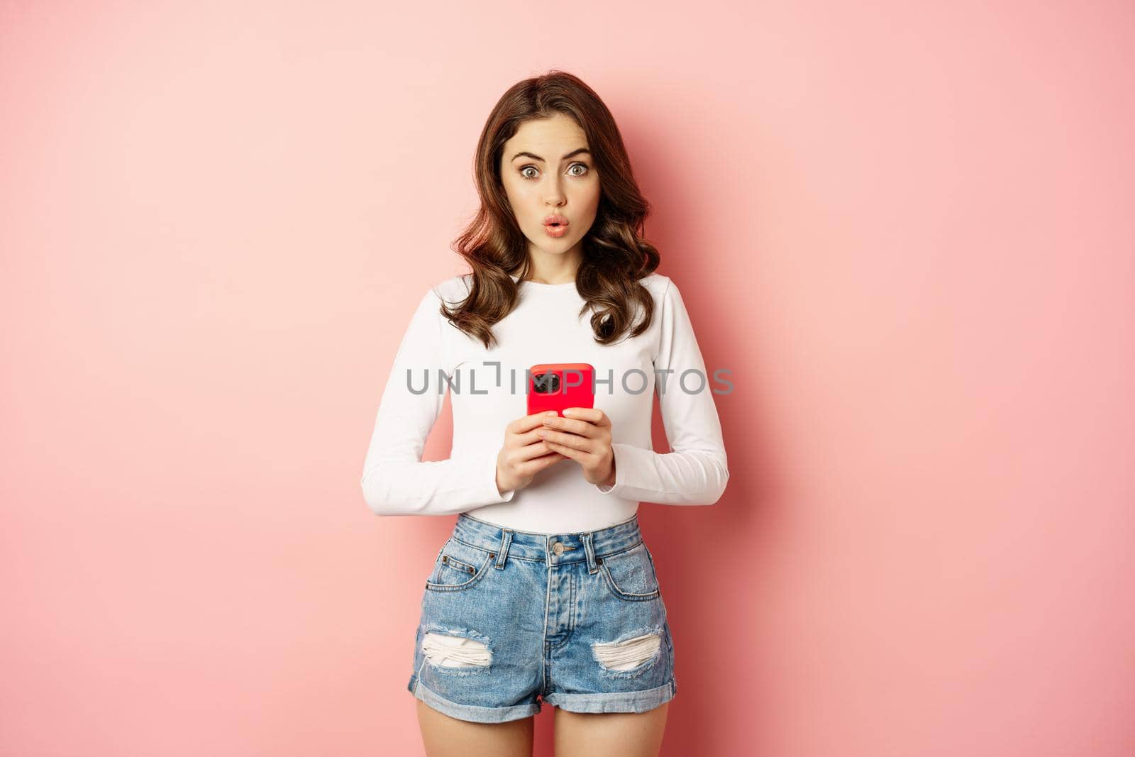 Surprised yong woman holding smartphone, looking amazed at camera, checking out advertisement, online shopping announcement, standing over pink background by Benzoix