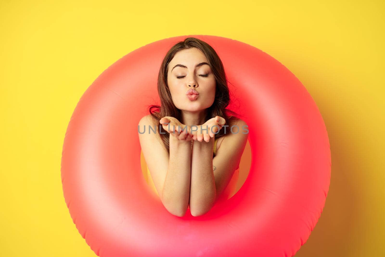 Portrait of beautiful female model posing with pink swimming beach ring, sending air kiss, mwah flirty, standing over yellow background by Benzoix