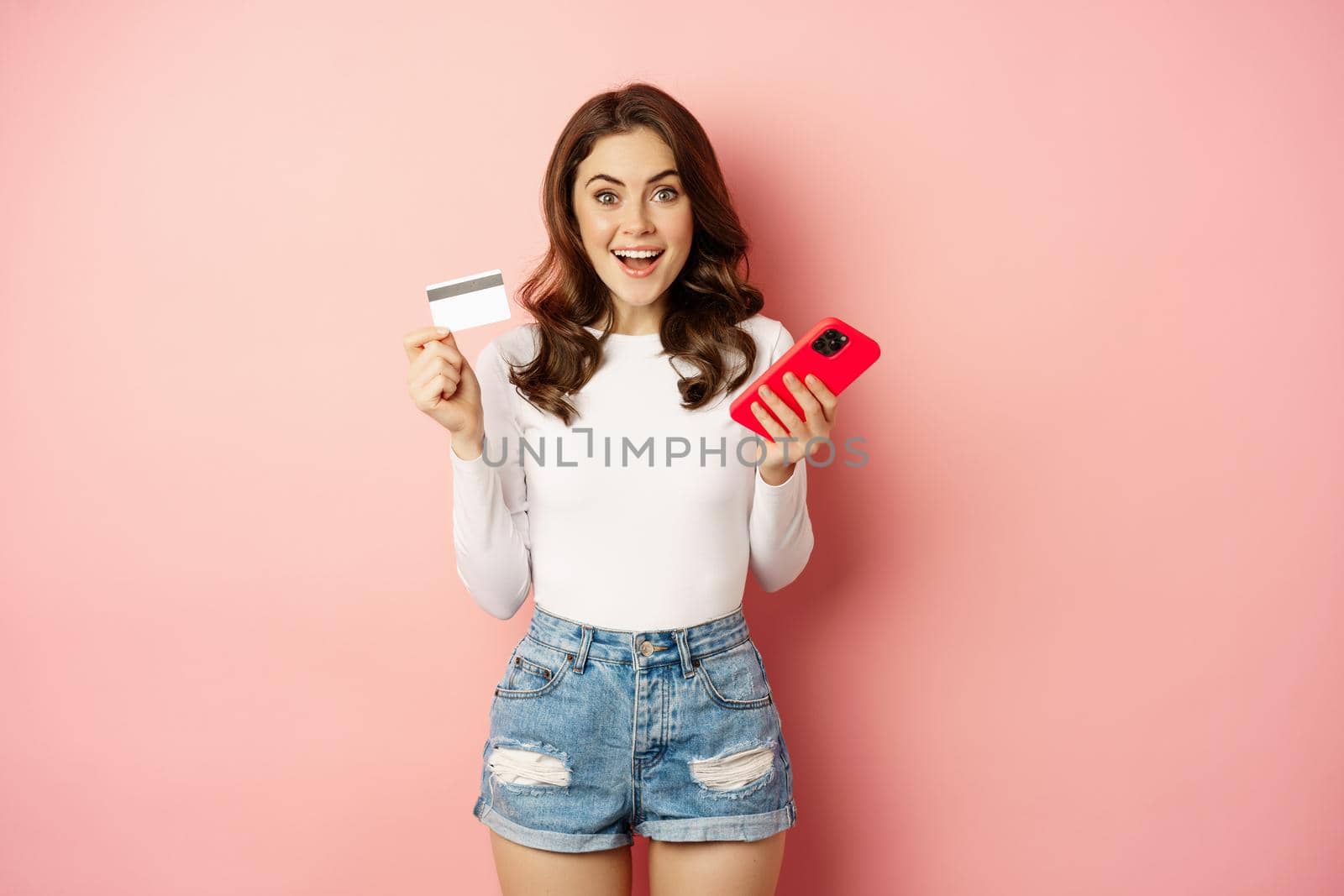 Enthusiastic brunette girl showing credit card and using mobile phone to order or pay, online shopping app, standing over pink background.