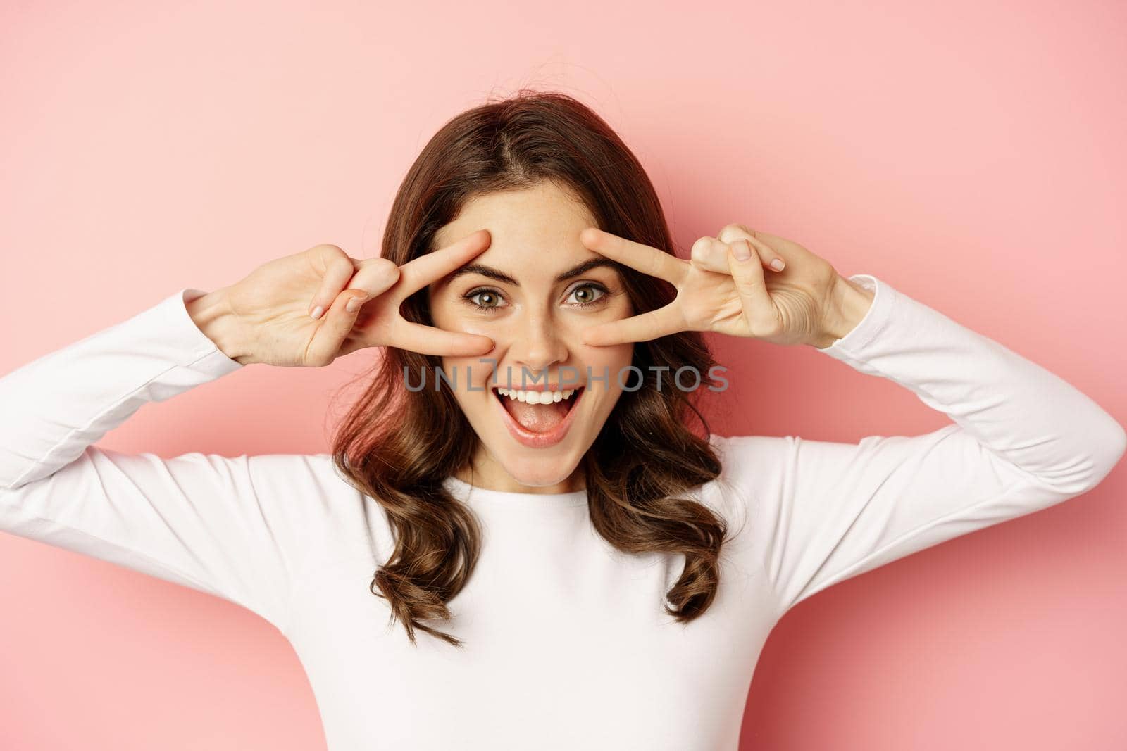Close up portrait of coquettish young woman smiling, showing peace, v-sign gesture and posing happy, standing against pink background.