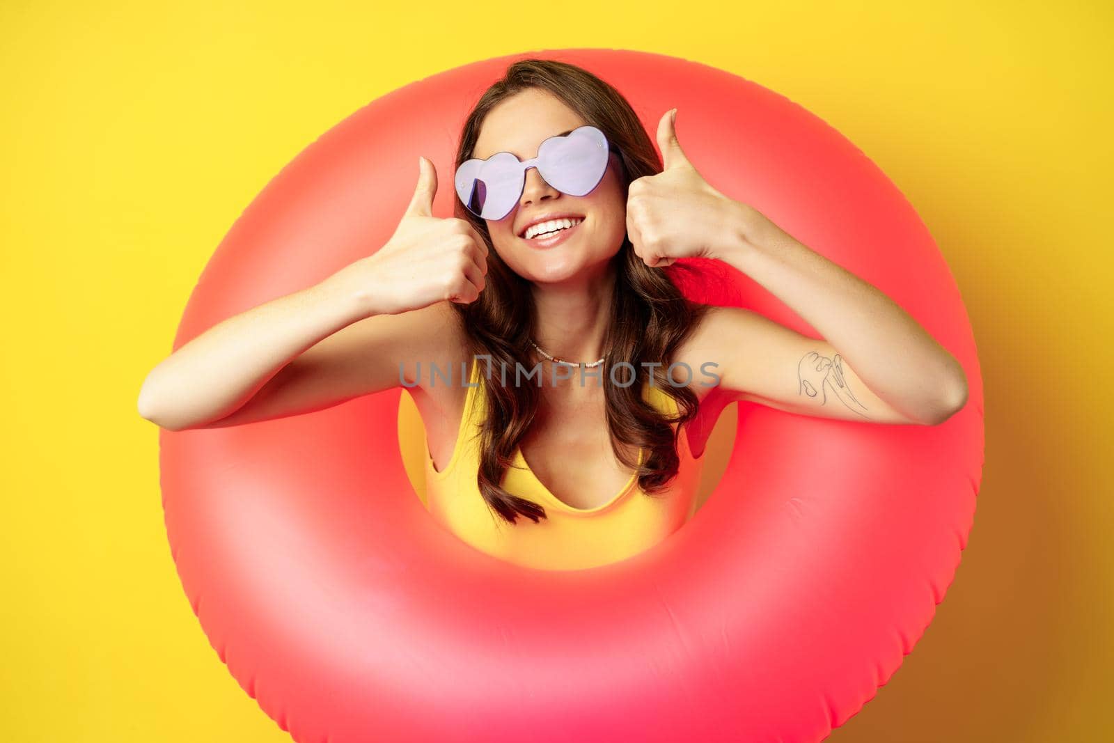 Close up portrait of happy attractive woman in sunglasses, wearing pink swimming ring, smiling and showing thumbs up, standing over yellow background.