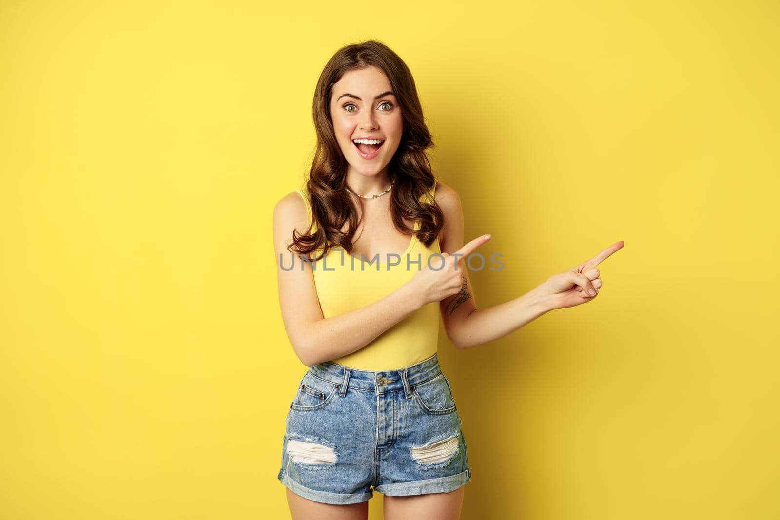 Beautiful young woman pointing fingers at banner, logo or advertisement, smiling amazed, standing in summer clothes, yellow background.