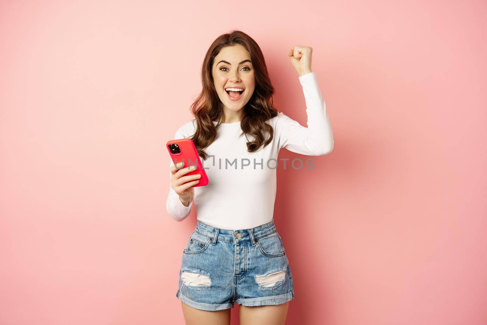 Enthusiastic brunette girl winning on mobile phone, holding smartphone and rejoicing, scream in joy, achieve app goal, pink background by Benzoix