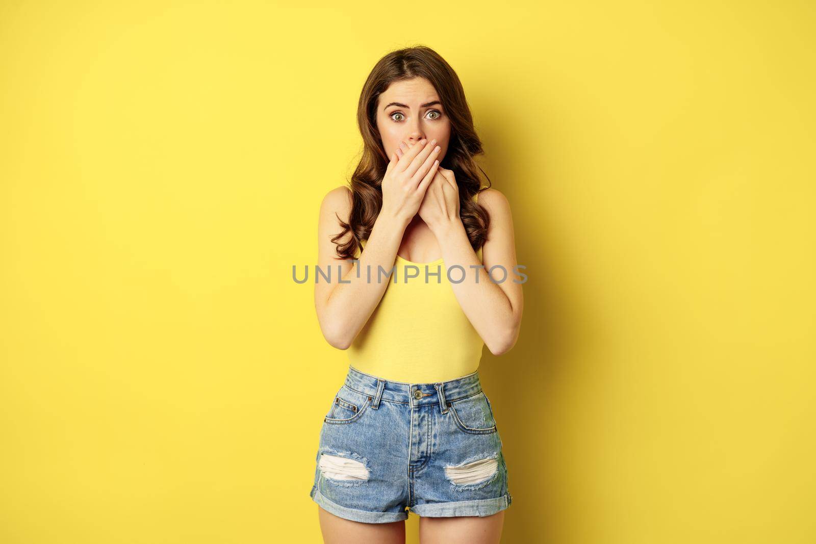 Shocked and scared young woman gasping, covering mouth with palms, looking frightened and startled, standing over yellow background.