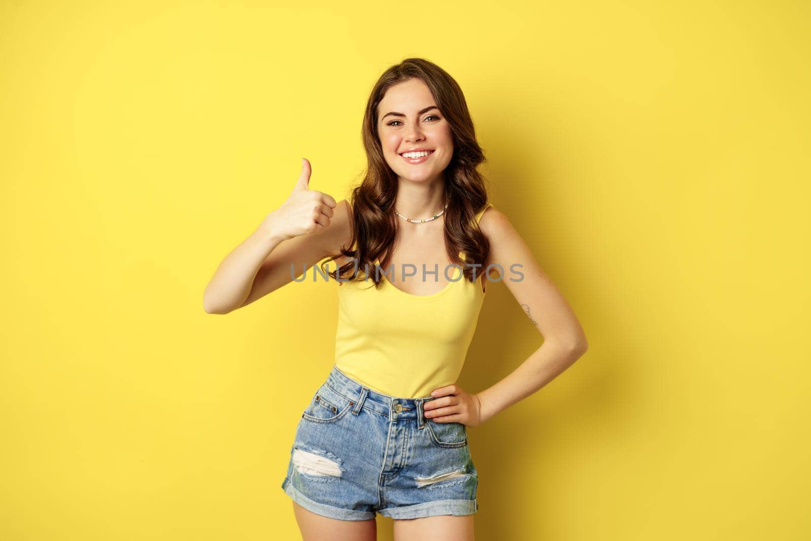 Portrait of stylish modern girl, feminine woman showing thumbs up, recommending gesture, like or approve, praise, standing over yellow background.