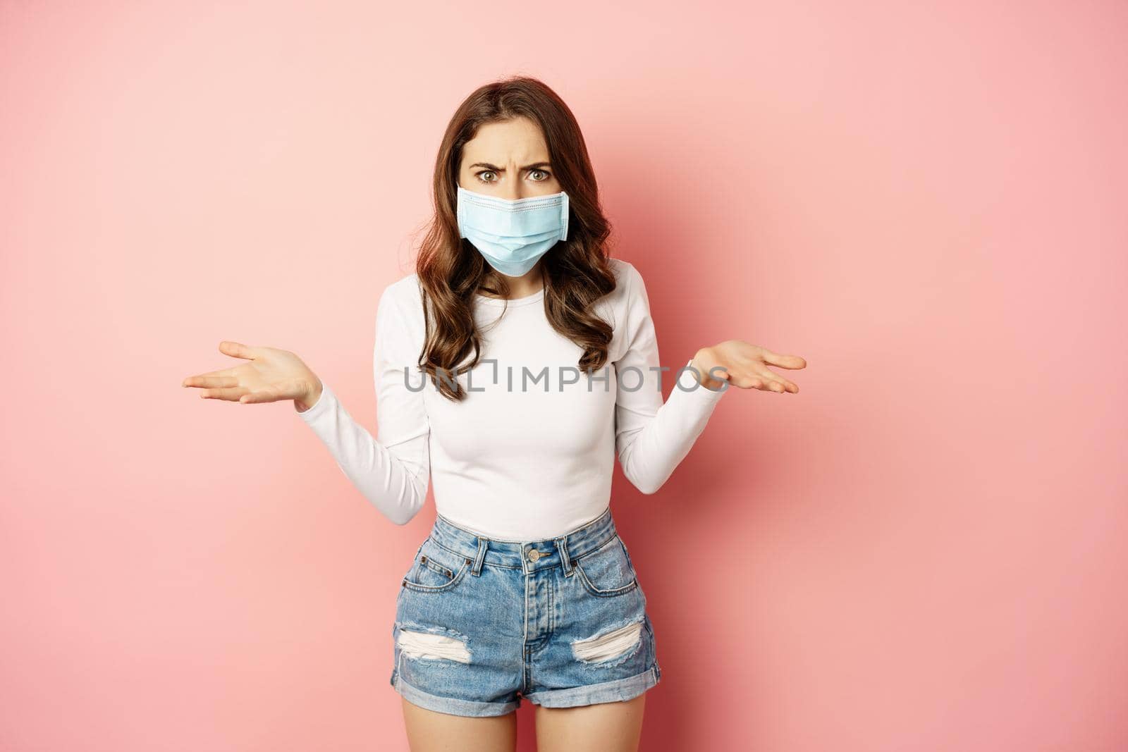 Covid-19. Confused young woman in medical face mask, shrugging shoulders and looking clueless, standing puzzled against pink background by Benzoix