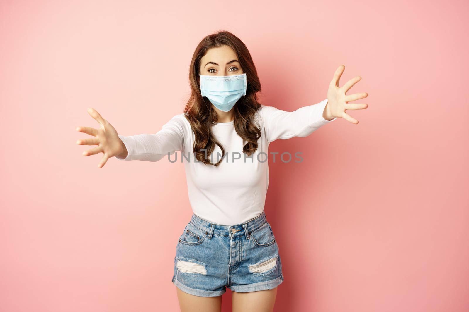 Portrait of smiling friendly girl in medical face mask, reaching hands, stretching arms forward for hug, receiving smth, standing over pink background by Benzoix