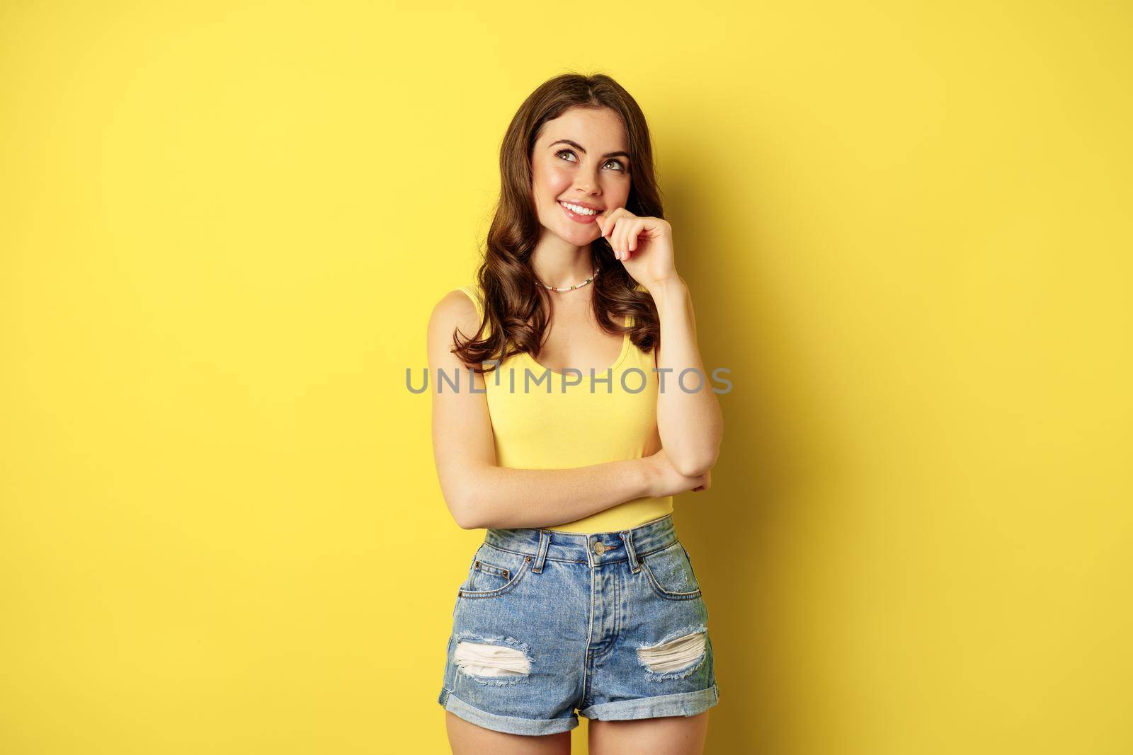 Thinking woman smiling, looking up and deciding, imaging smth, picturing or daydreaming, posing in summer clothes against yellow background.