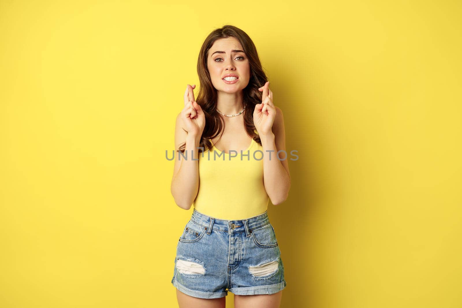 Hopeful young beautiful woman praying, believe, hoping to receive smth, cross fingers for good luck, anticipating, standing over yellow background.