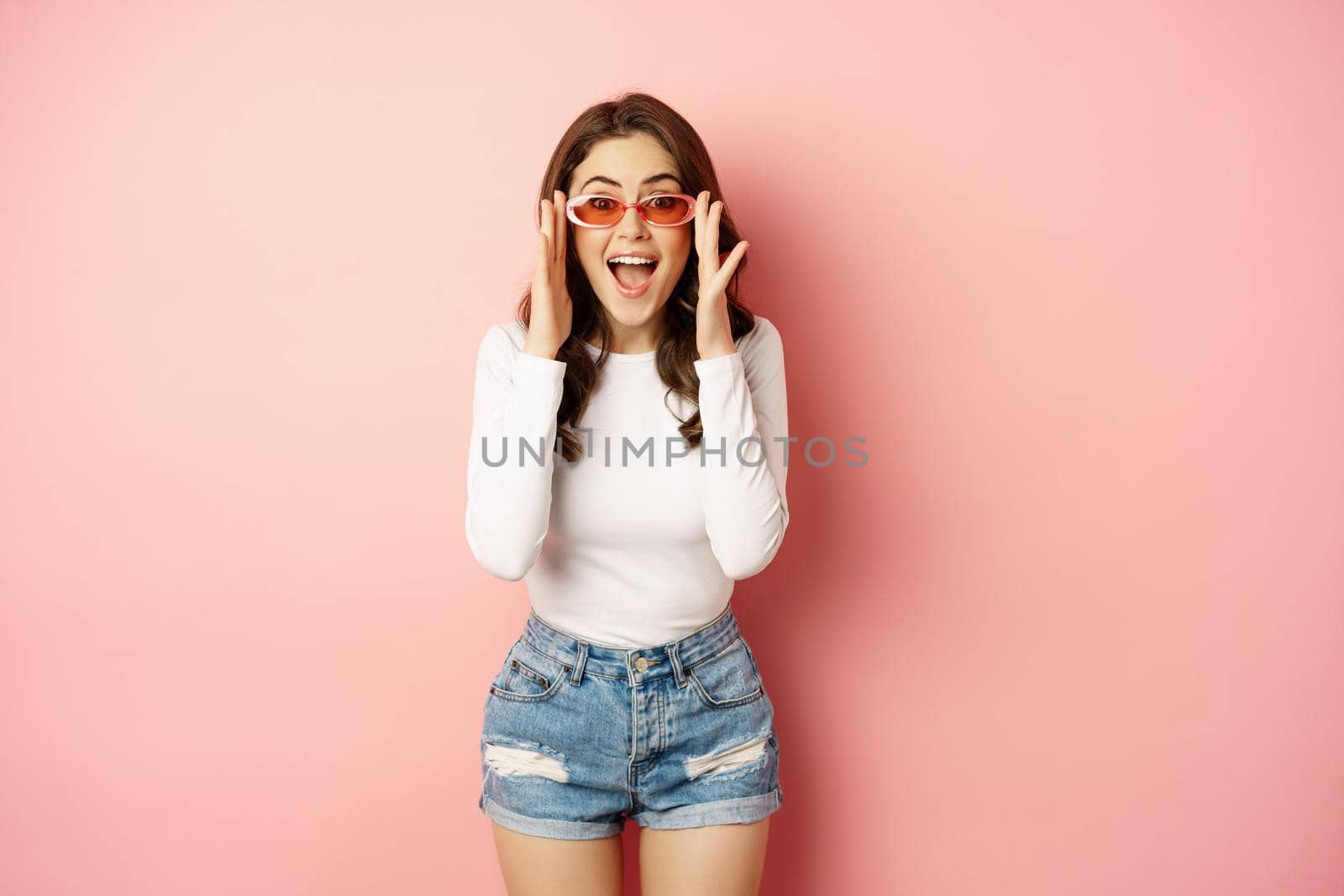 Enthusiastic brunette girl in stylish sunglasses, reacts surprised and amazed, looking with disbelief at camera, standing over pink background.