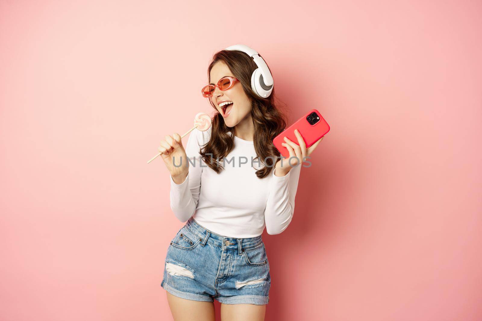 Summer girl laughing, eathing lolipop and listening music in headphones, dancing with smartphone against pink background.