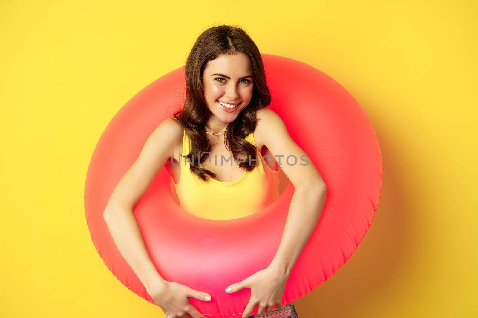 Happy beautiful young woman relaxing on summer vacation, wearing pink swim ring for beach holiday, standing against yellow background.