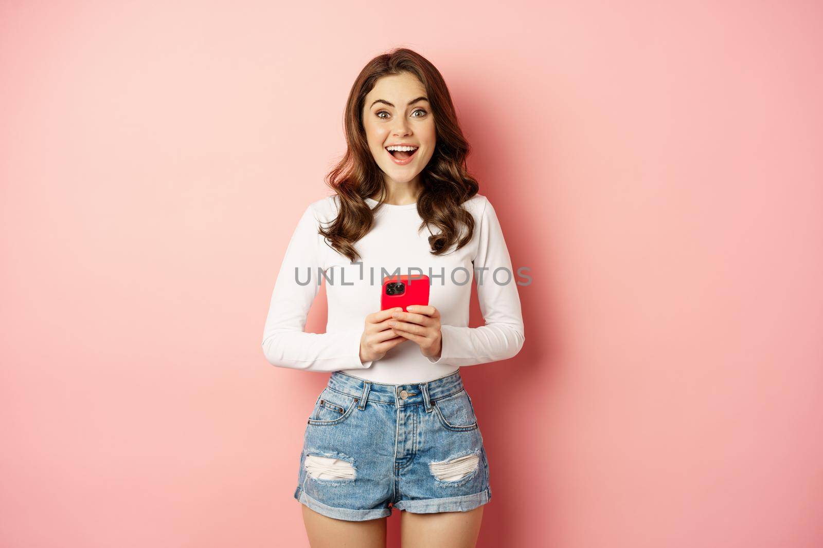 Online shopping and cellphone concept. Excited attractive girl holding mobile phone and smiling, standing over pink background.