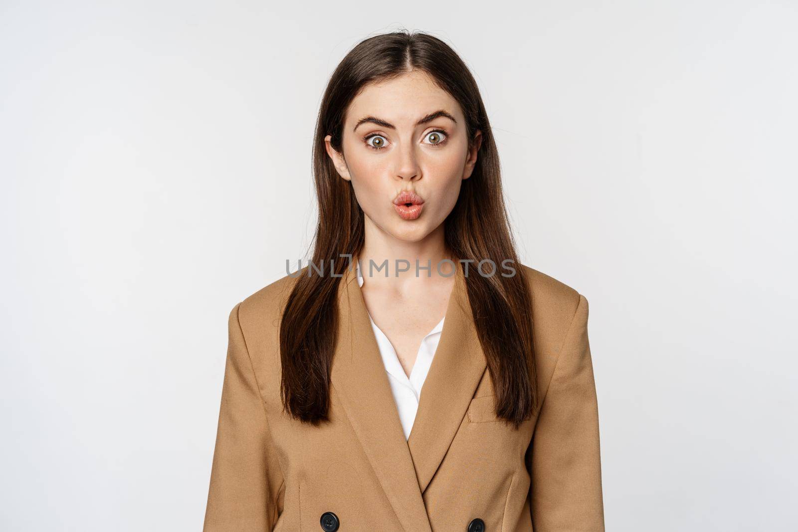 Portrait of office lady at workplace, looking surprised and curious, wow face, standing over white background in brown suit.