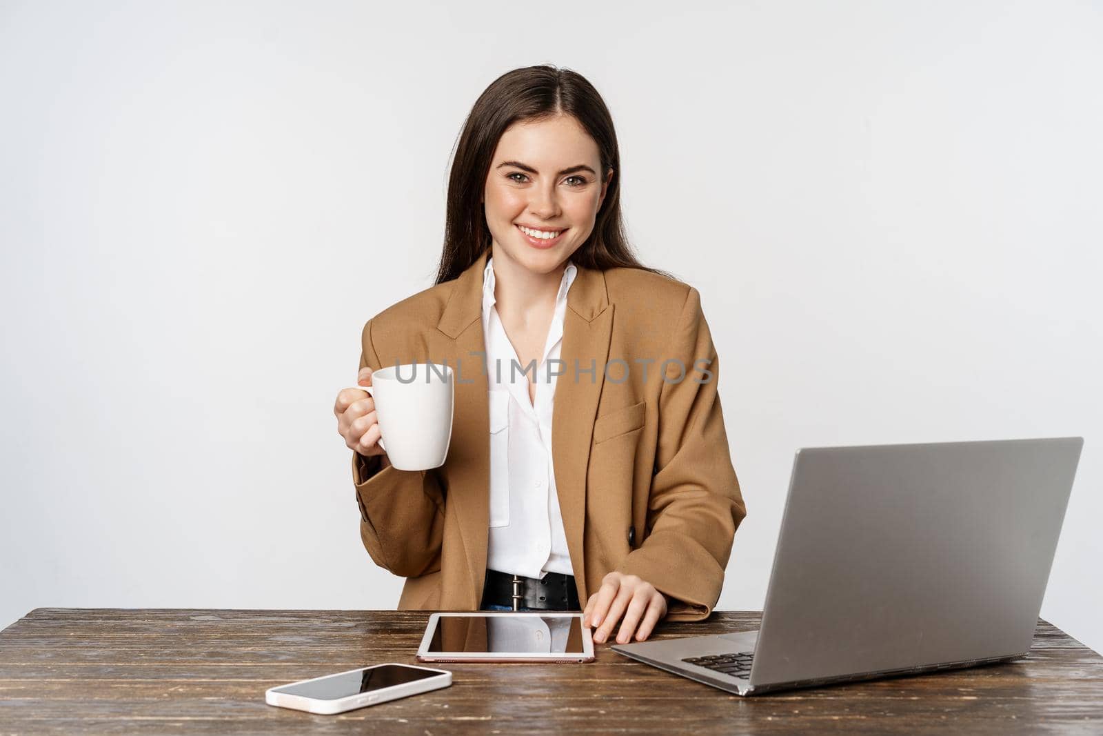 Beautiful happy office lady, business woman drinking coffee at work, sitting at table and smiling pleased, holding white mug, studio background.