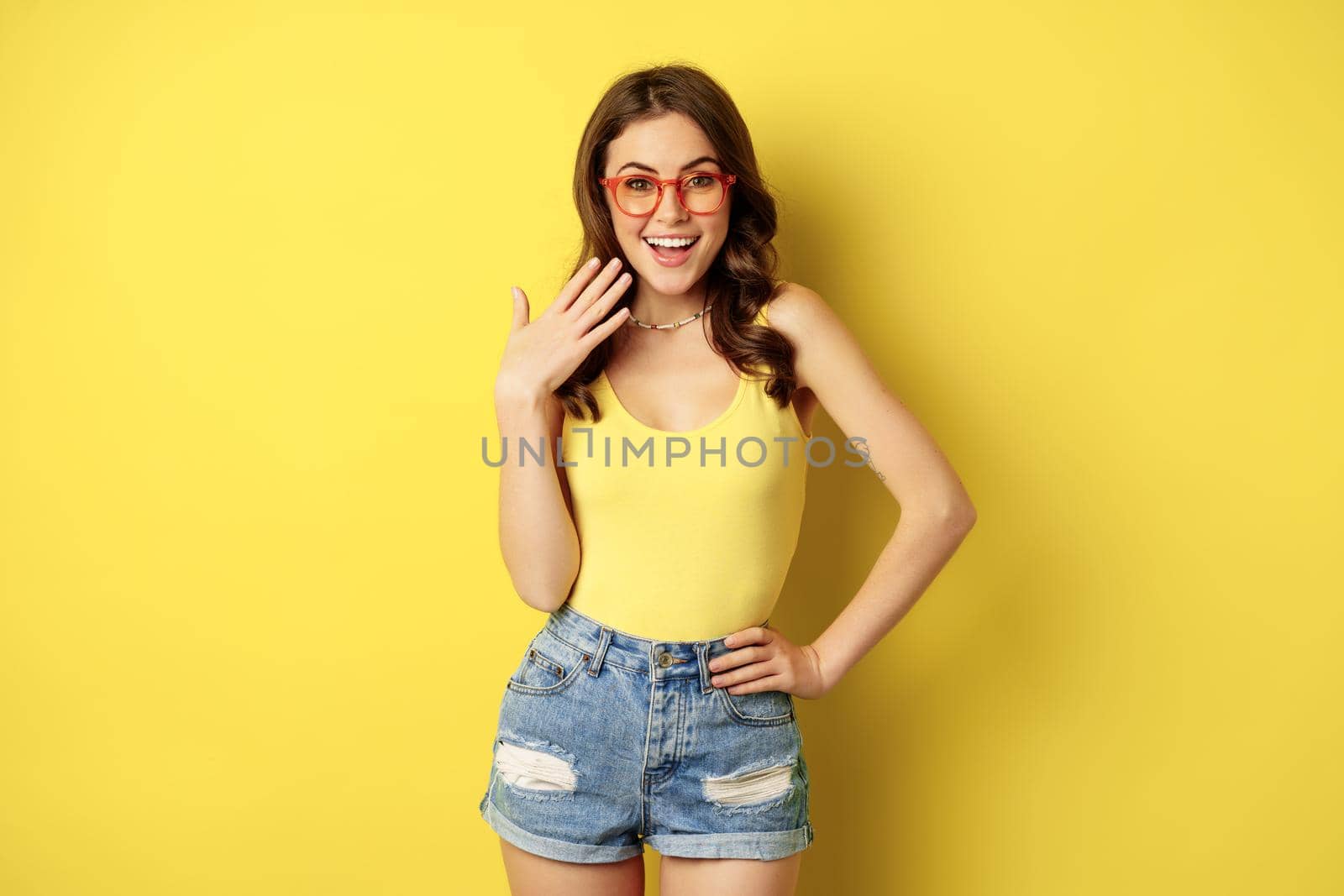 Coquettish girl in sunglasses, laughing and smiling, waving hand at herself, reacting to smth amazing, standing over yellow background.