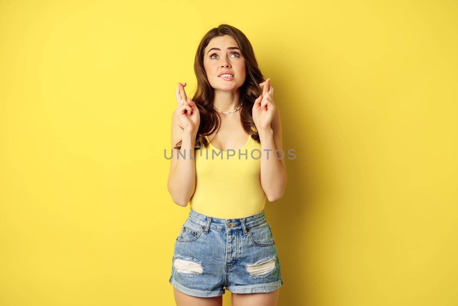 Hopeful young beautiful woman praying, believe, hoping to receive smth, cross fingers for good luck, anticipating, standing over yellow background by Benzoix