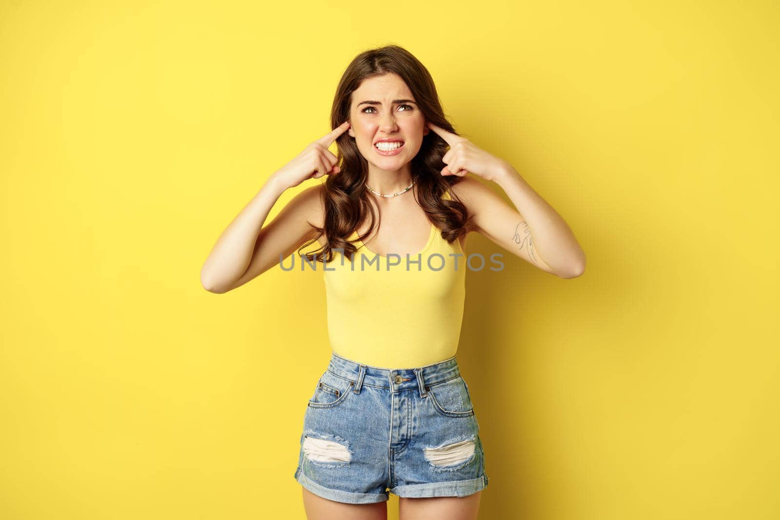 Young stylish woman feeling discomfort from loud noise sound, shut ears with fingers and complaining, standing over yellow background by Benzoix