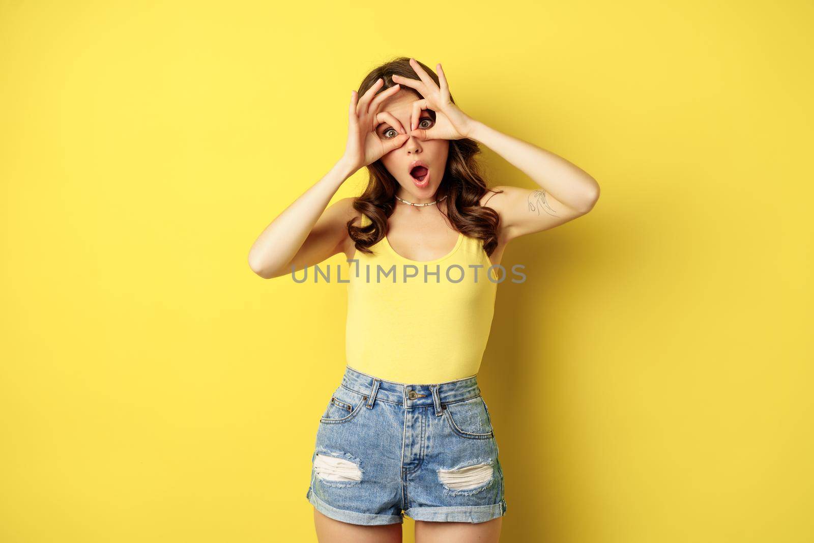 Positive young woman showing okay, ok zero signs, face mask gesture, grimacing and having fun, posing in tank top against yellow background by Benzoix