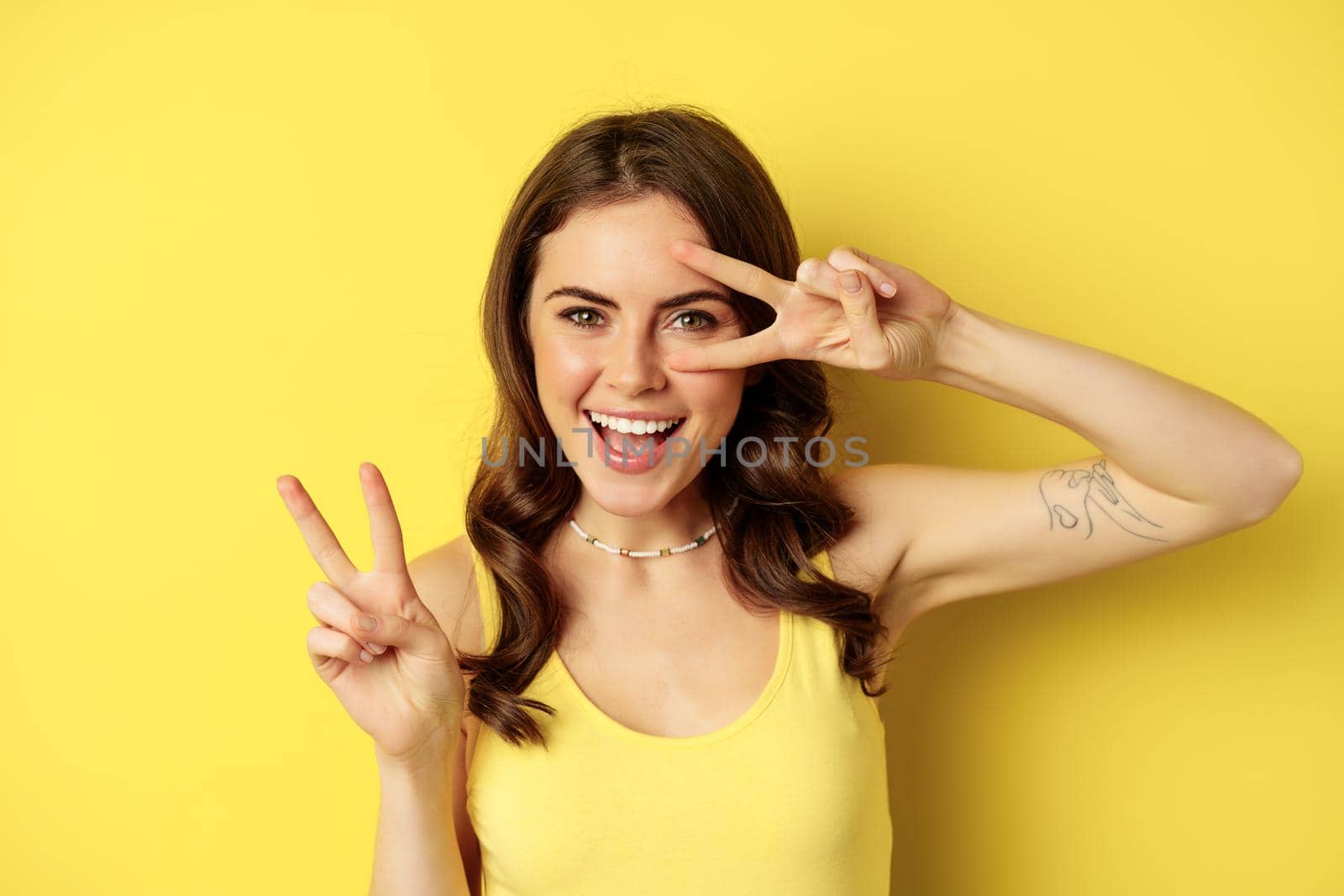 Close up portrait of stylish happy girl, showing peace sign and smiling, posing against yellow background. Copy space