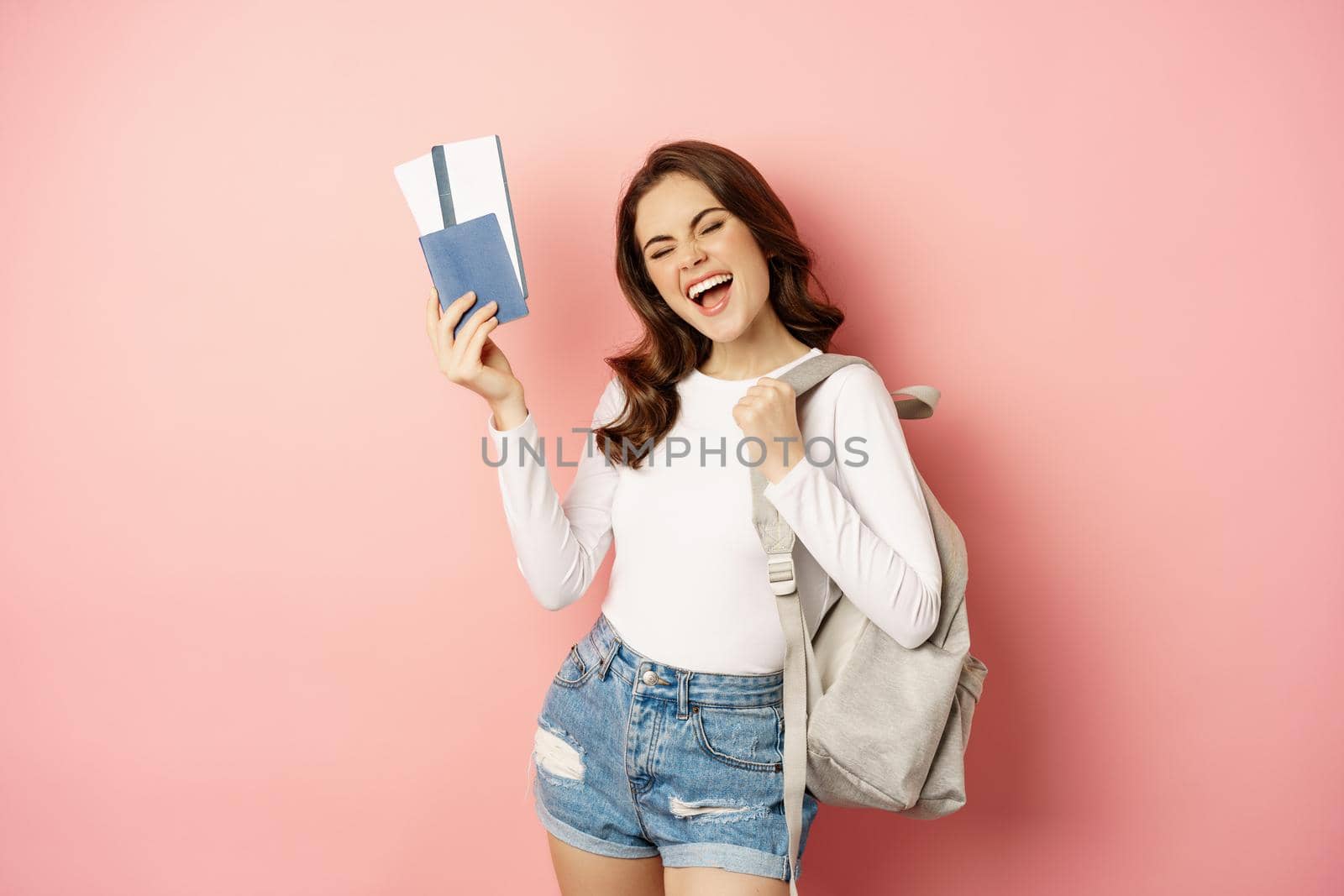 Vacation and holiday. Beautiful girl going on trip, holding passport with airplane tickets, holding backpack, travelling, standing over pink background.