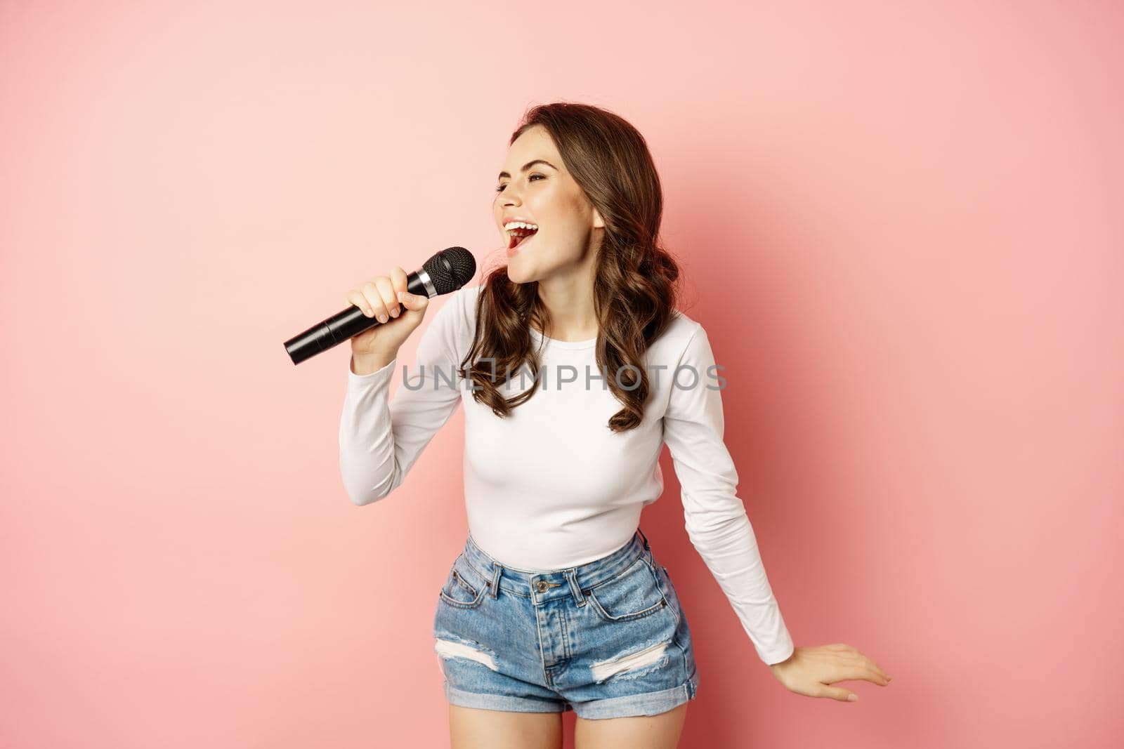 Party girl. Happy young woman singing in microphone, performing song, having fun at event, standing over pink background by Benzoix