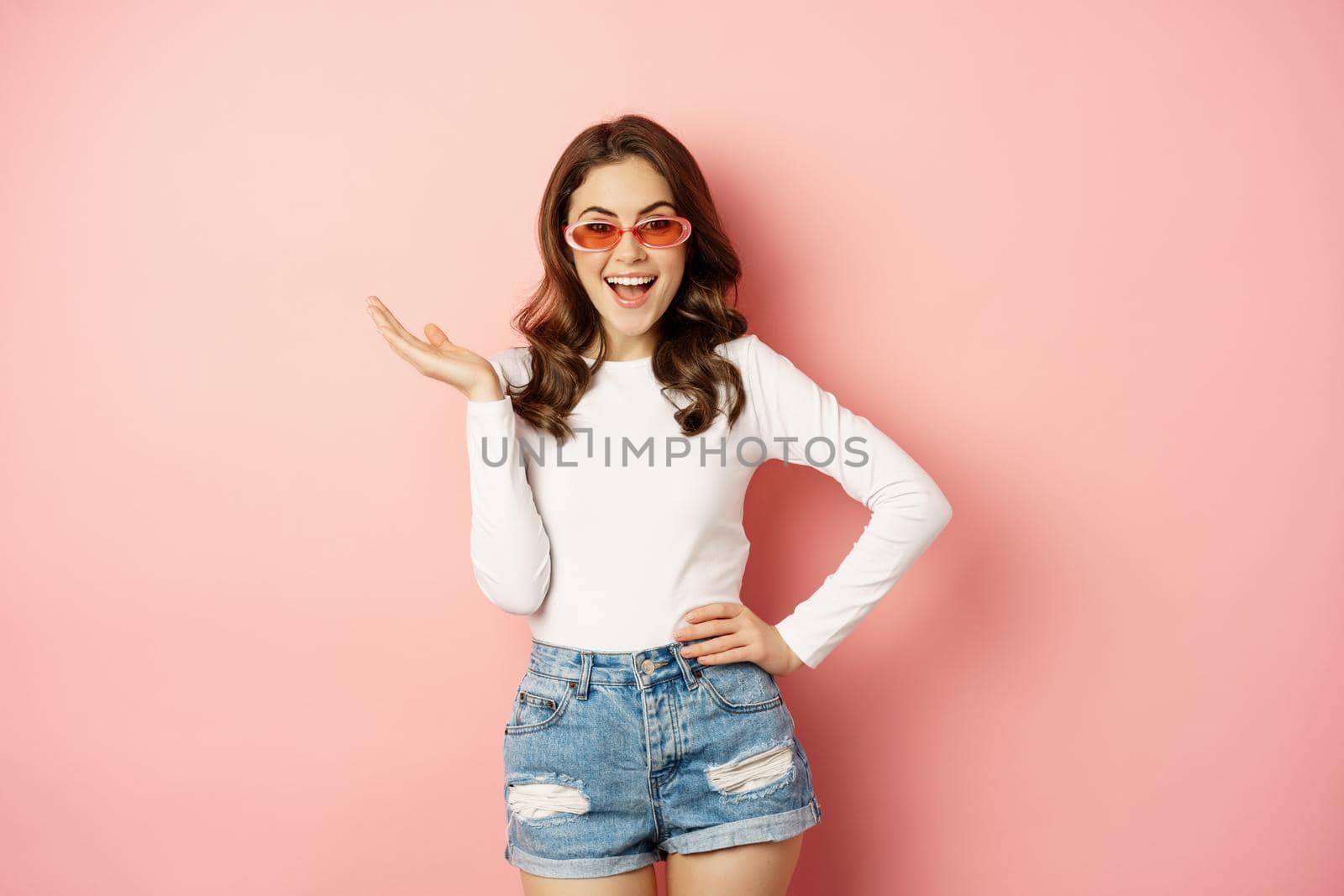Coquettish and stylish brunette girl, laughing and smiling, wearing sunglasses, wearing white blouse and jeans, pink background. Copy space