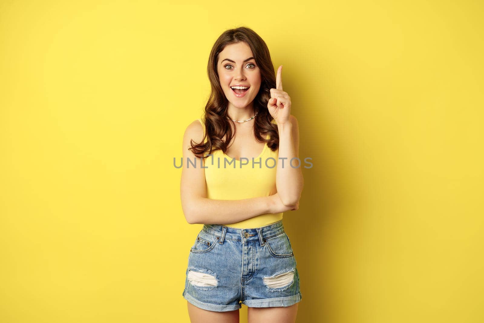 Creative young woman has suggestion, raising finger, eureka sign, has an idea, standing in summer outfit over yellow background by Benzoix