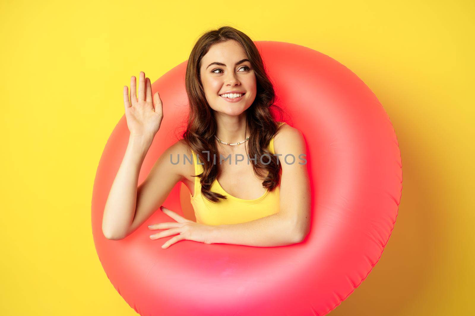 Happy young woman wearing pink swimming ring, waving hand and saying hi, enjoying vacation, summer beach holiday, standing over yellow background.