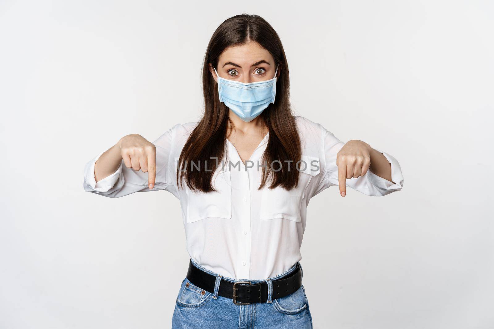 Covid and people concept. Young woman in face mask pointing fingers down, showing advertisement, company logo below, standing over white background.