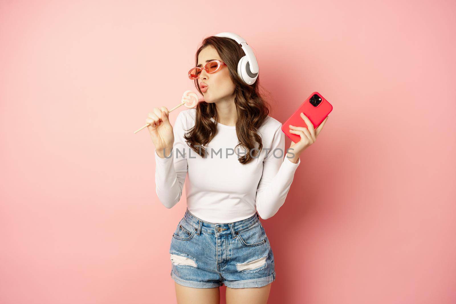 Summer girl laughing, eathing lolipop and listening music in headphones, dancing with smartphone against pink background.