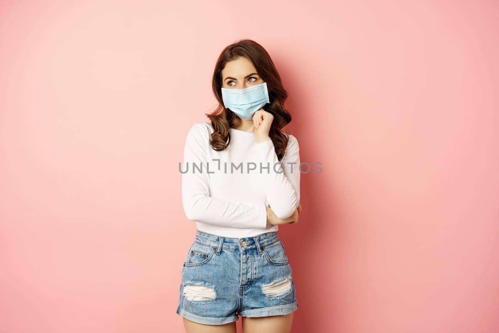 Portrait of stylish modern girl in medical face mask, looking coquettish and smiling at camera, standing over pink background. Pandemic and covid concept.