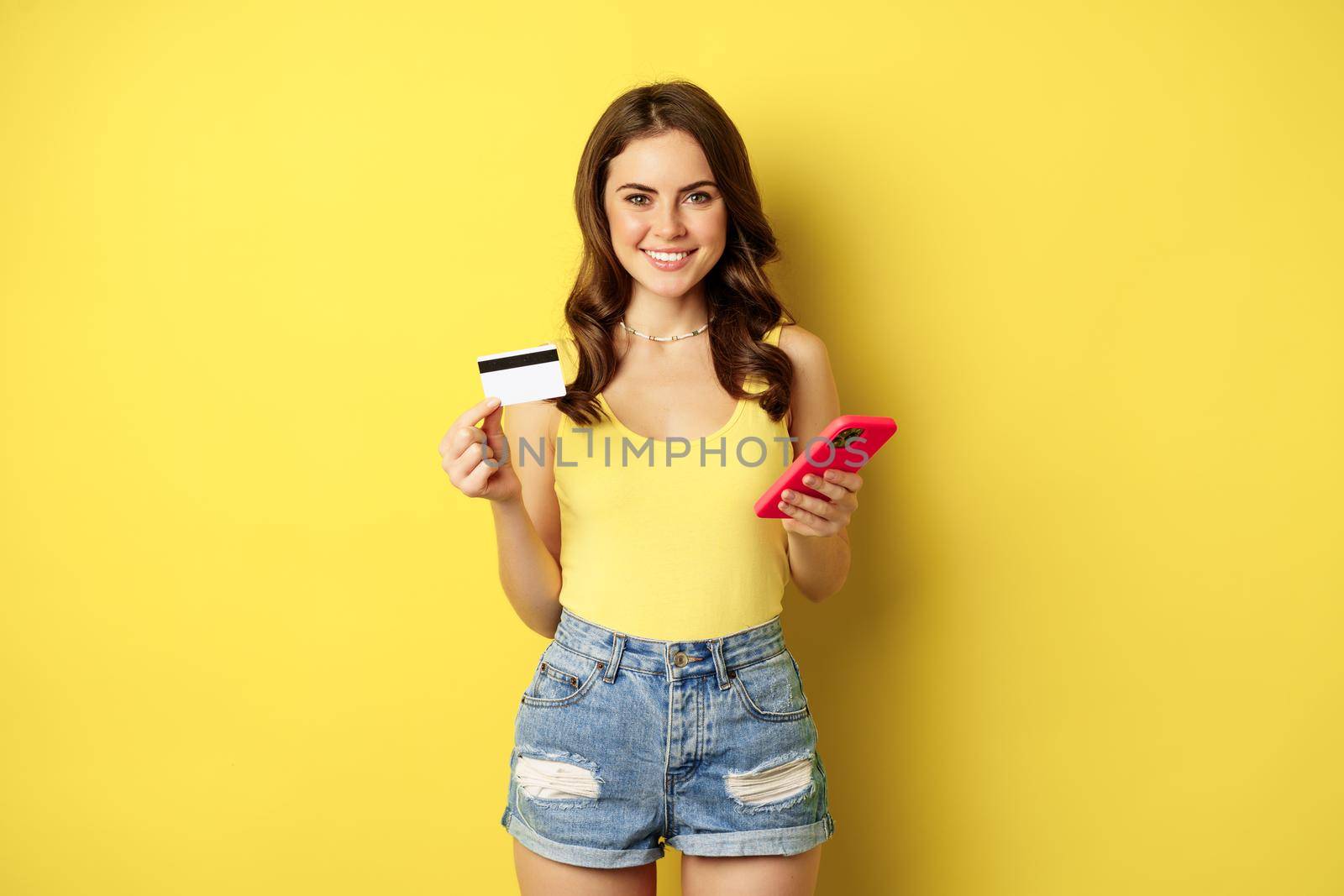 Stylish young brunette woman, holding credit card and smartphone, paying, purchase or order something in store, standing over yellow background by Benzoix