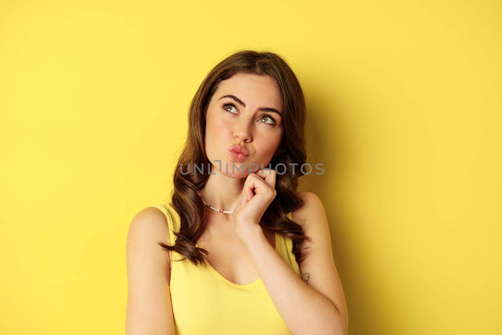 Close up portrait of stylish young beautiful woman thinking, yearning smth, looking up and making choice, standing over yellow background.