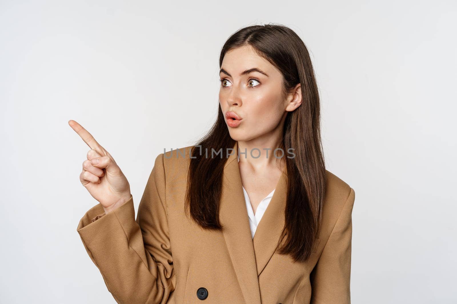 Portrait of businesswoman pointing finger left, showing corporate banner, logo, standing in brown suit over white background.