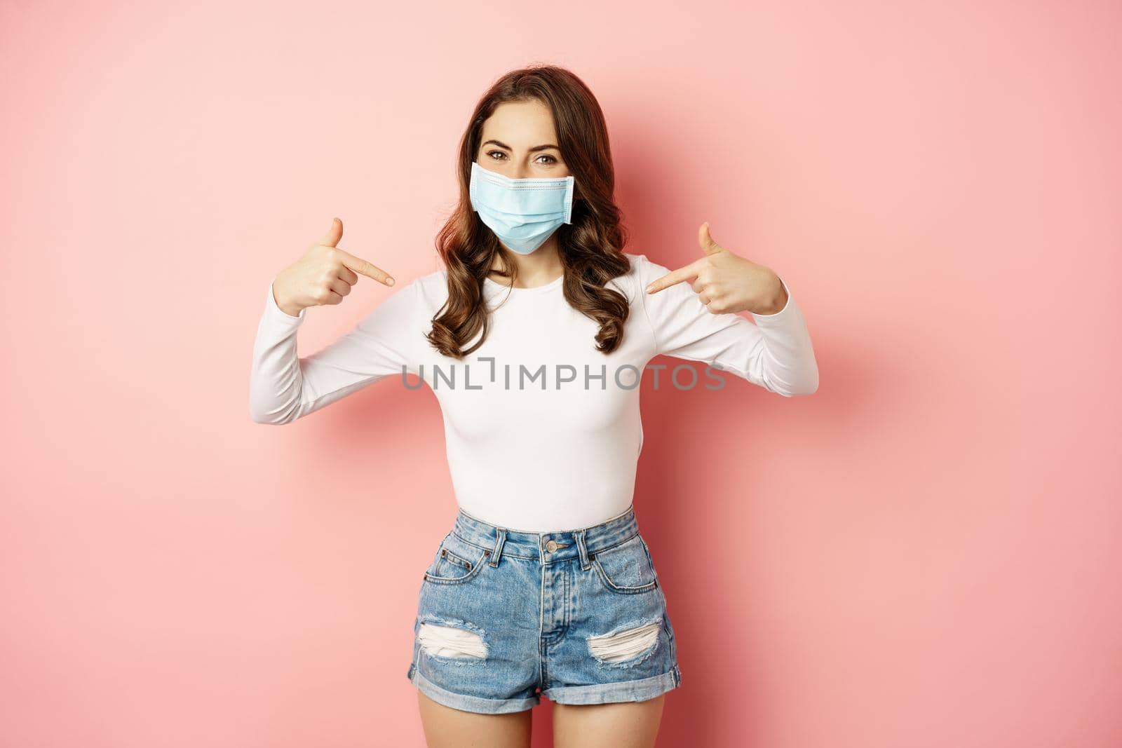 Young woman in medical face mask, pointing fingers at herself, showing logo or banner, smiling at camera, standing over pink background by Benzoix