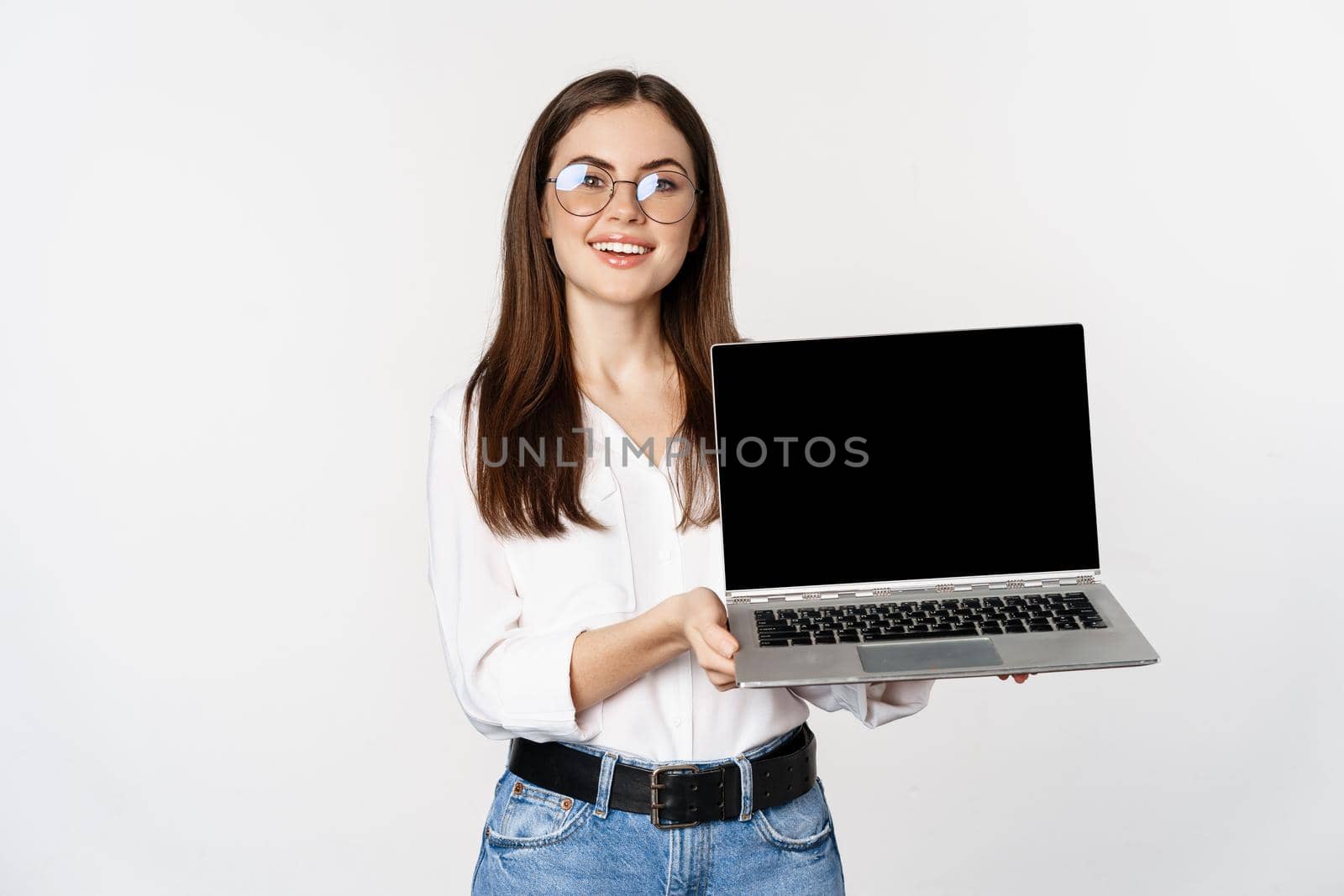 Young woman in glasses showing laptop screen, demonstrating promo on computer, website or store, standing over white background.