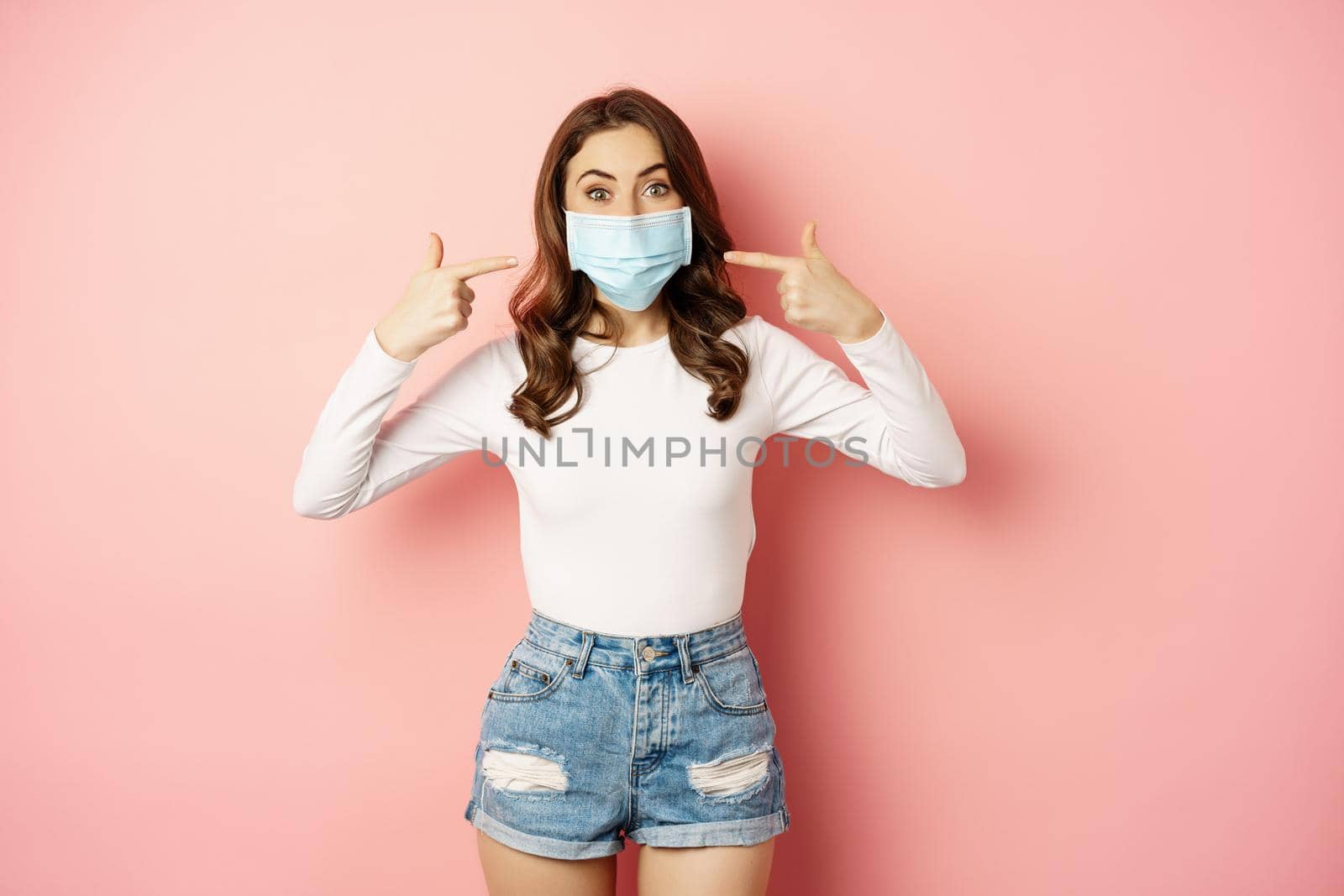 Portrait of young stylish woman wearing and pointing fingers at her medical face mask, smiling, preventive measures during covid 19 pandemic, quarantine, standing over pink background by Benzoix